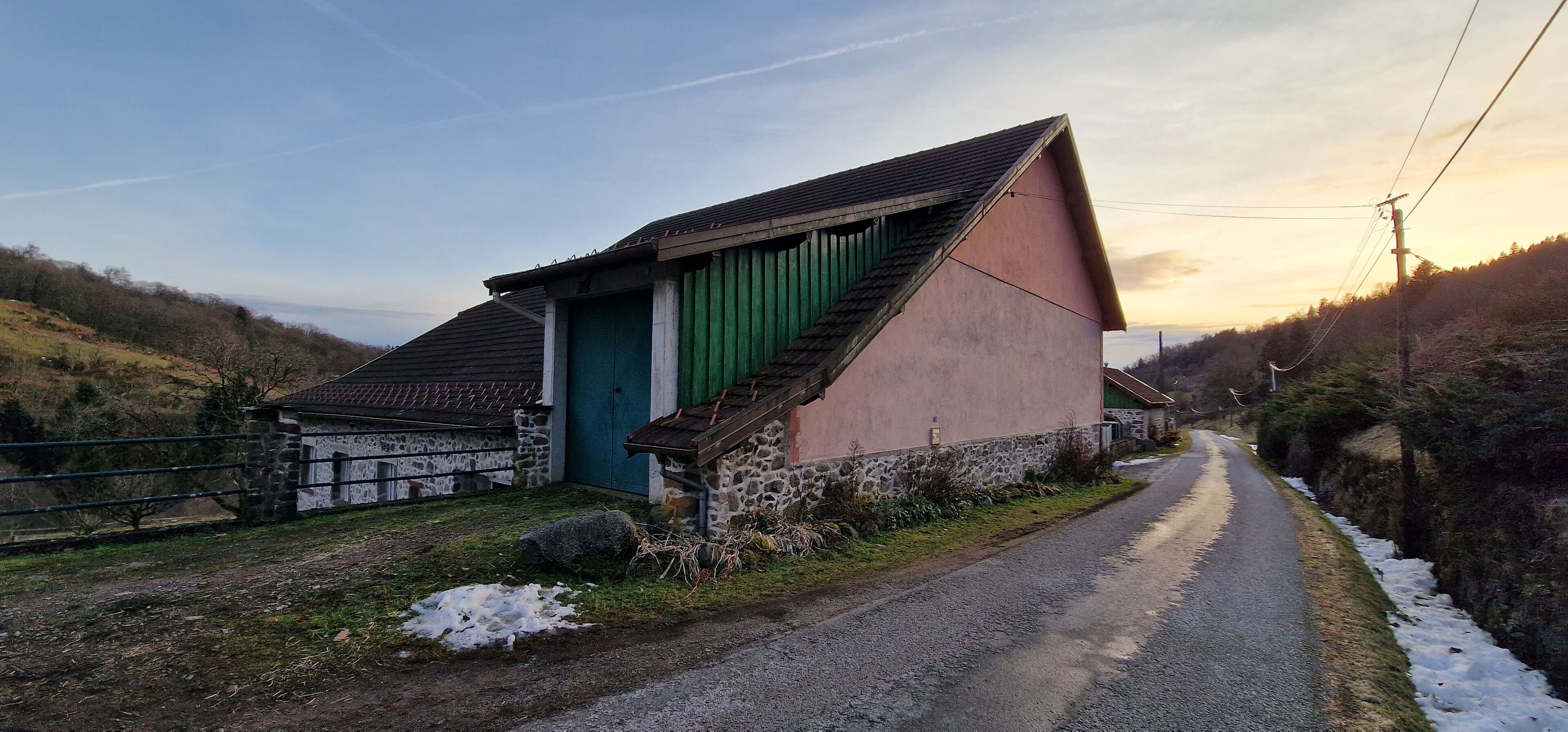 Una strada di campagna con una casa in pietra a sinistra, un portone verde e un muro rosa.