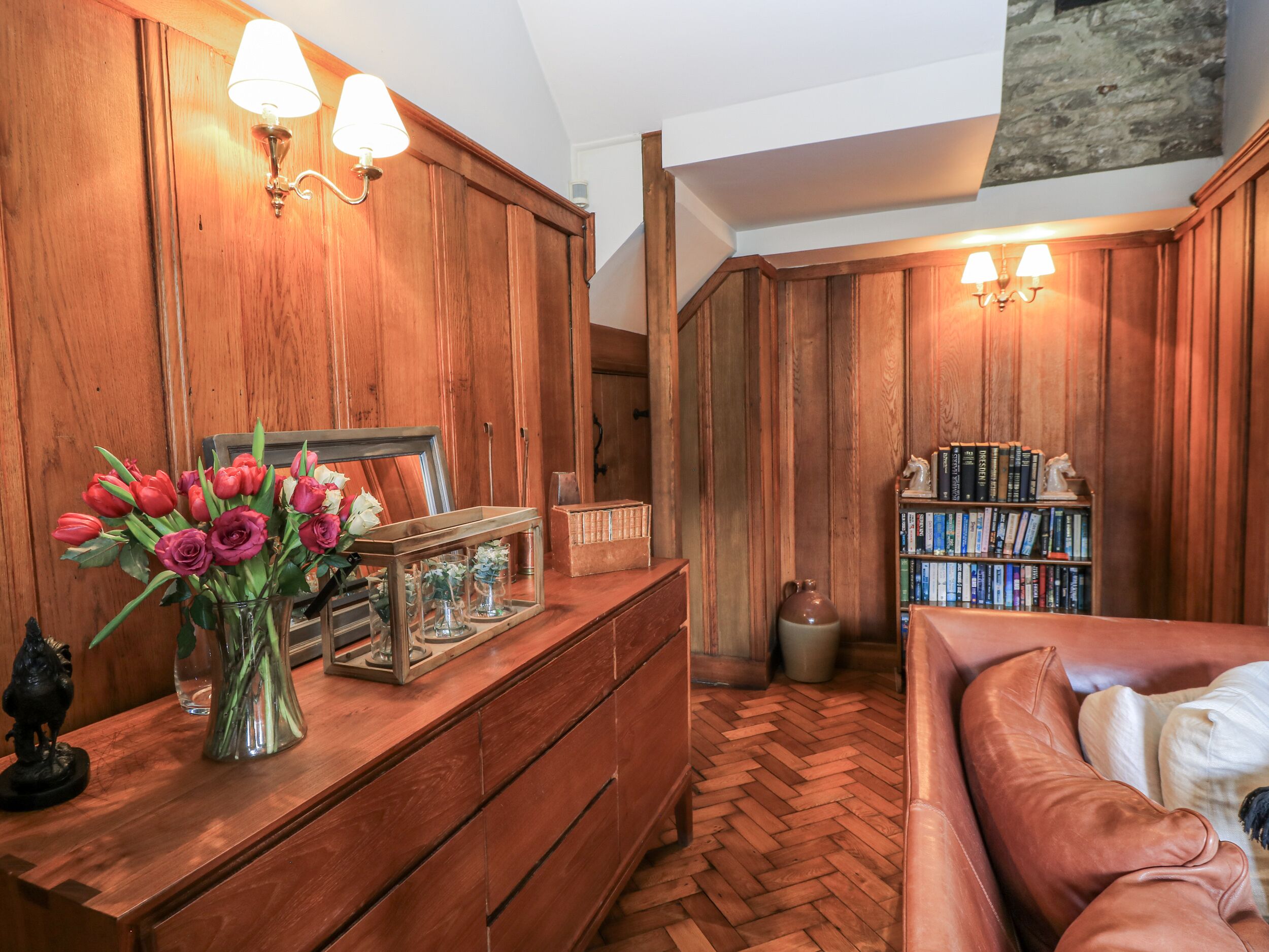 A room with wooden paneling, a herringbone floor, a wooden dresser with a mirror, a vase of flowers, a bookshelf, and a leather couch.