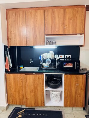 A compact kitchen with wooden cabinets, a black countertop, a microwave, and a sink. - 1Bedroom 1Bath | Perfect for Couples & Business (Nairobi)