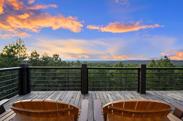 Una terraza de madera con dos sillas y una barandilla, con vista a un paisaje boscoso bajo un cielo de atardecer.