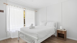 A minimalist bedroom with a white bed, a wooden nightstand, and sheer curtains. - The Harla Beachside - ideally situated near the azure ocean, pristine beaches just a short stroll (Coffs Harbour)