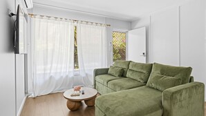 A green sofa, a round wooden coffee table, and a white air conditioning unit in a room with white walls and a wooden floor. - The Harla Beachside - ideally situated near the azure ocean, pristine beaches just a short stroll (Coffs Harbour)