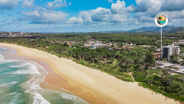 A coastal view with a beach, buildings, and a logo for 'COFFS HOLIDAY PROFESSIONALS'. - The Harla Beachside - ideally situated near the azure ocean, pristine beaches just a short stroll (Coffs Harbour)