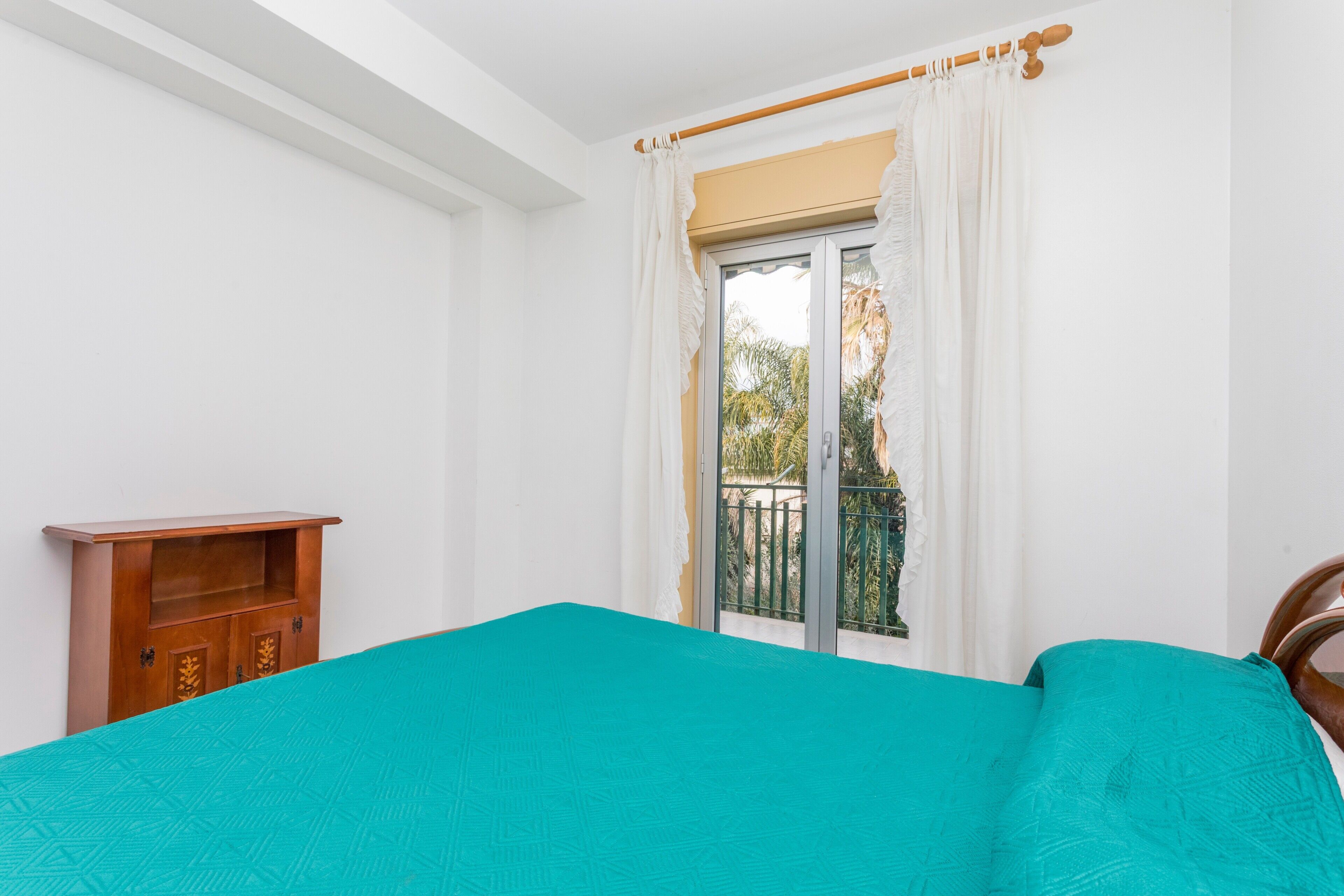 A room with a bed, a wooden cabinet, a window with curtains, and a balcony.