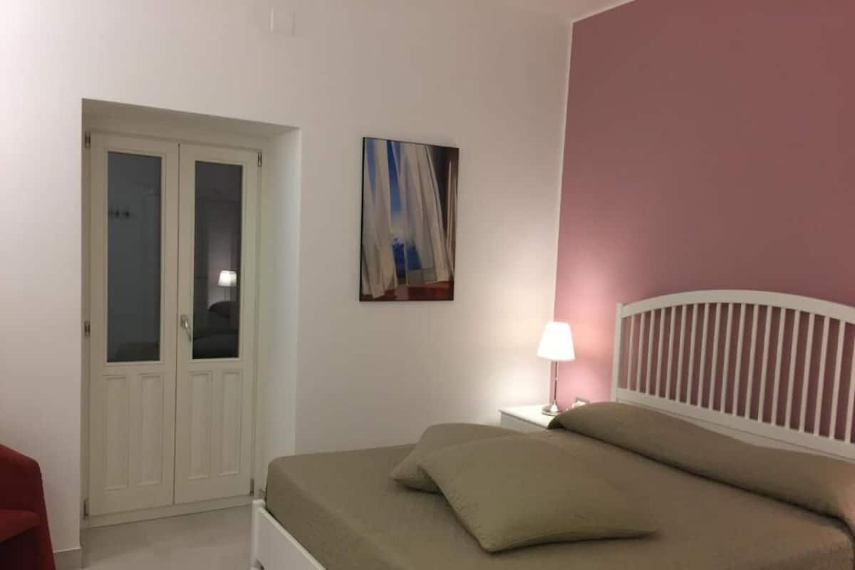 Quarto com cama, mesa de cabeceira, abajur, porta e um quadro na parede.