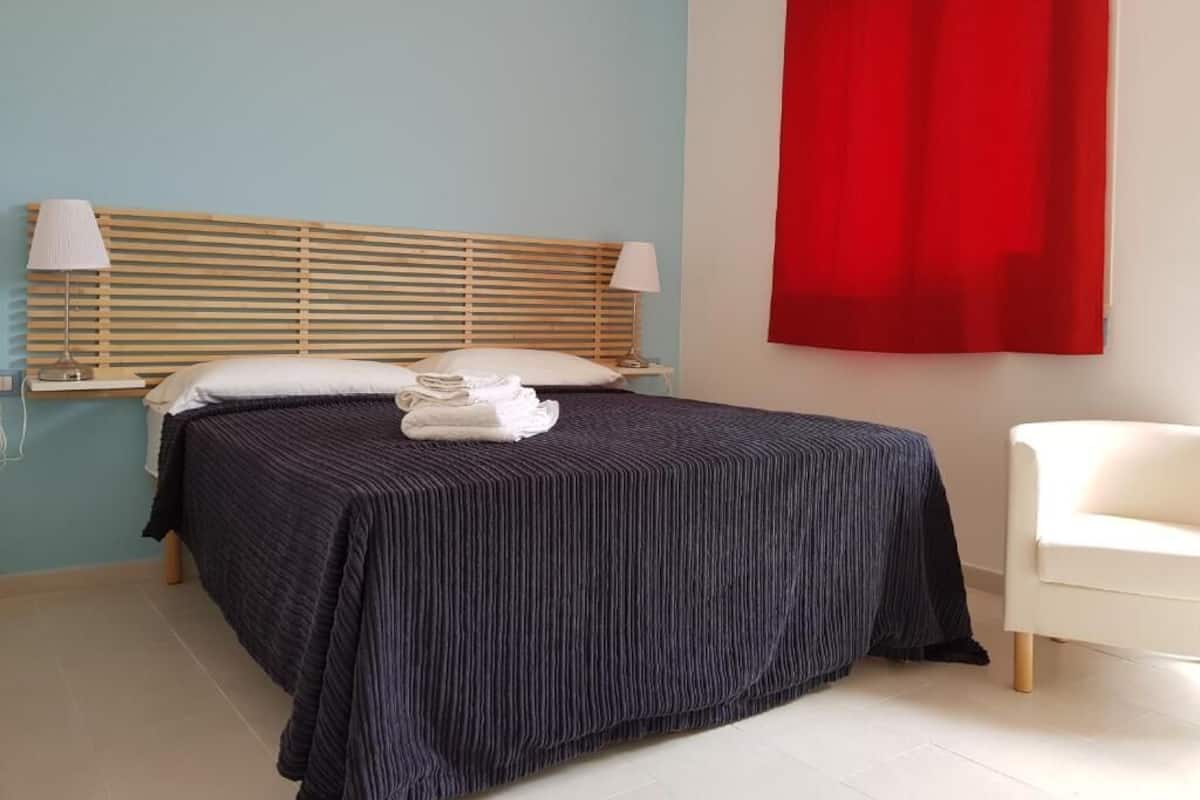 Cama bem arrumada com colcha listrada, duas luminárias de cabeceira, uma cortina vermelha e uma cadeira branca num quarto com paredes azul claro.