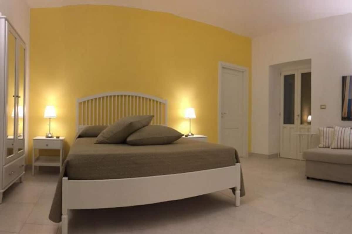 Quarto de hotel com cama, mesas de cabeceira, sofá e um quadro na parede.