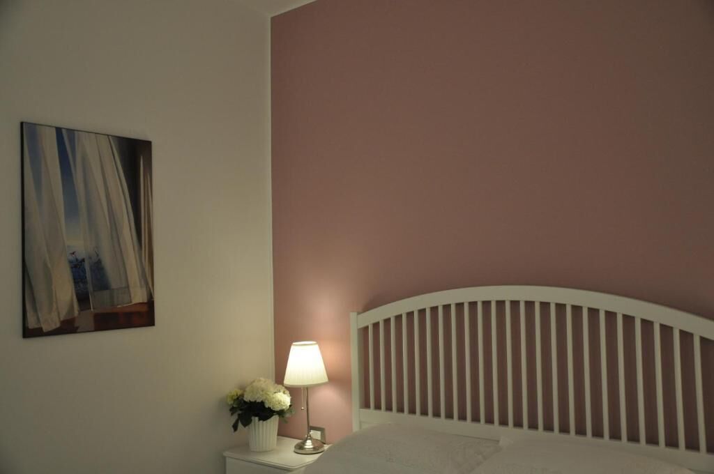 A bedroom with a white headboard, a bedside table with a lamp and flowers, and a painting on the wall.