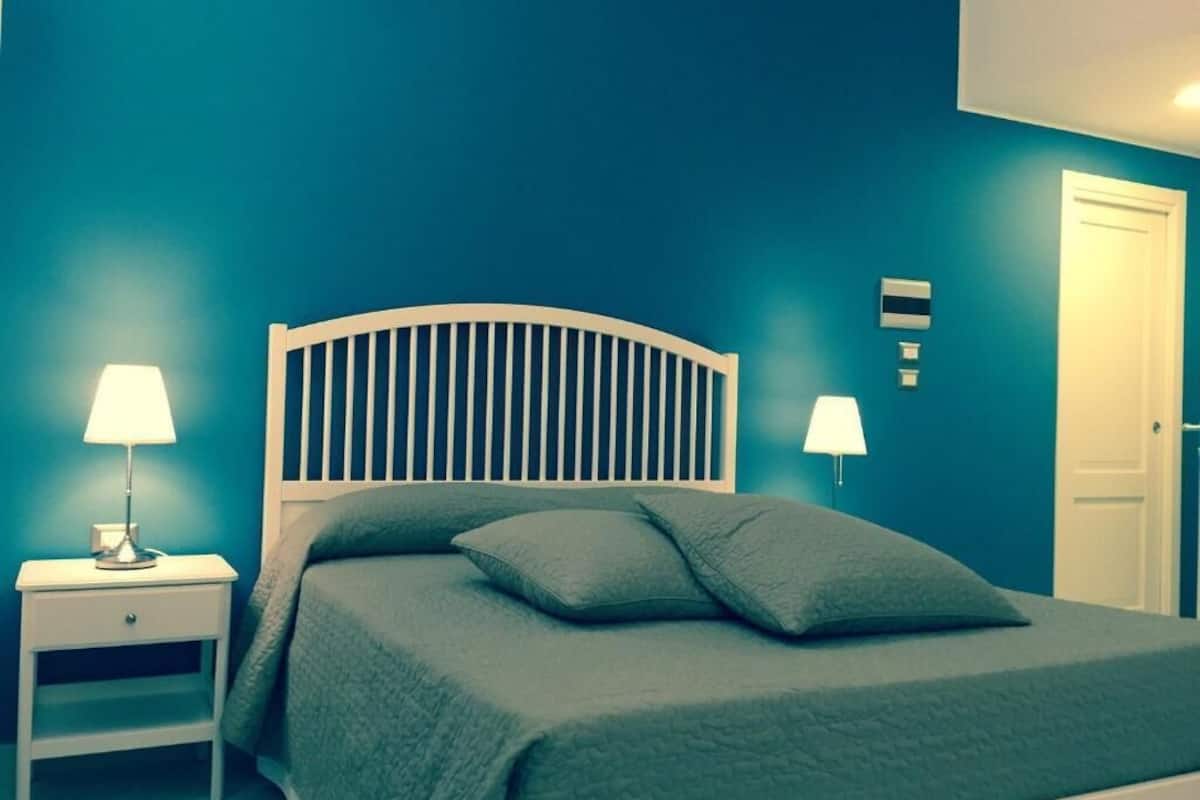Um quarto com uma cama, duas luminárias de cabeceira, uma mesinha de cabeceira e uma porta.