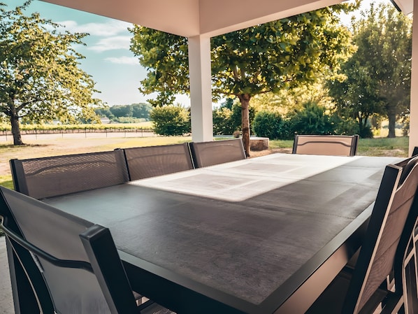 Un espacio de estar al aire libre cubierto, con una mesa y sillas, rodeado de árboles y vistas a un viñedo.