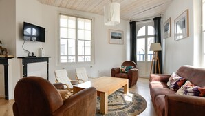 Uma sala de estar com mesa de centro de madeira, dois sofás de couro marrom, uma poltrona branca, uma TV fixada na parede e janelas amplas com cortinas.