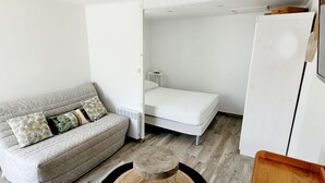 Een compacte kamer met een grijze bank, een wit matras, een klein tafeltje en een aan de muur bevestigde televisie.