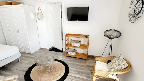 Een slaapkamer met een bed, een kledingkast, een televisie, een rieten stoel en een klein rond tafeltje met een geweven vloerkleed.