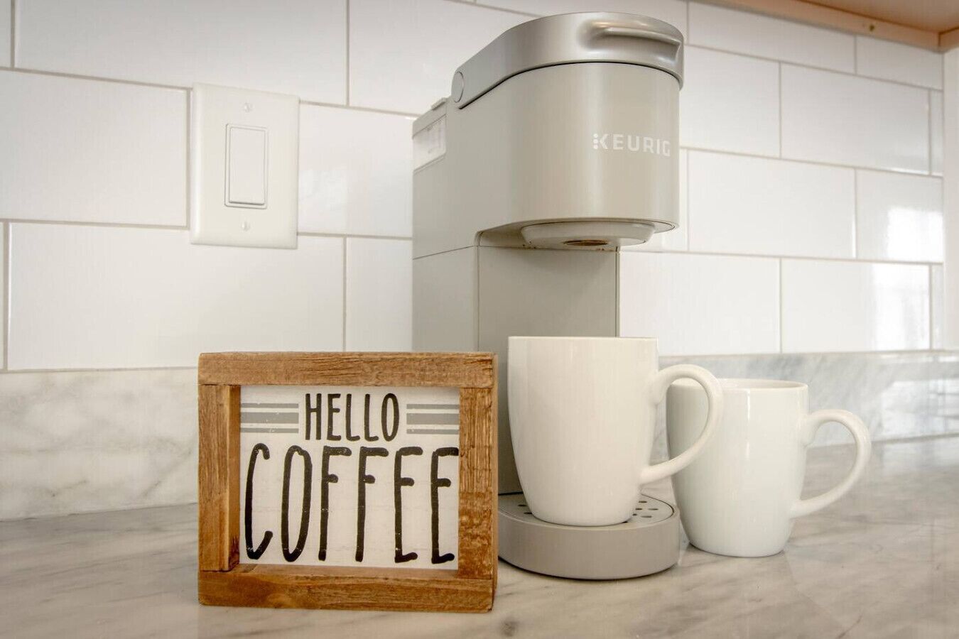 Une machine à café, deux tasses blanches et un panneau en bois sur lequel on peut lire « HELLO COFFEE », le tout posé sur un plan de travail de cuisine.