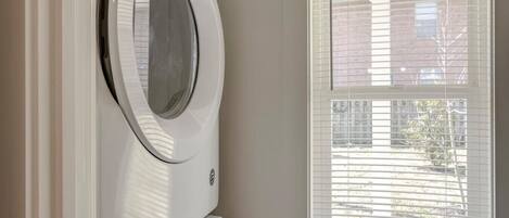 A laundry room with a front-loading washer and dryer, a window with blinds, and a view of a building outside.