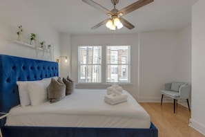 A bedroom with a tufted blue headboard, a large bed with white linens, a ceiling fan, and a window with blinds.