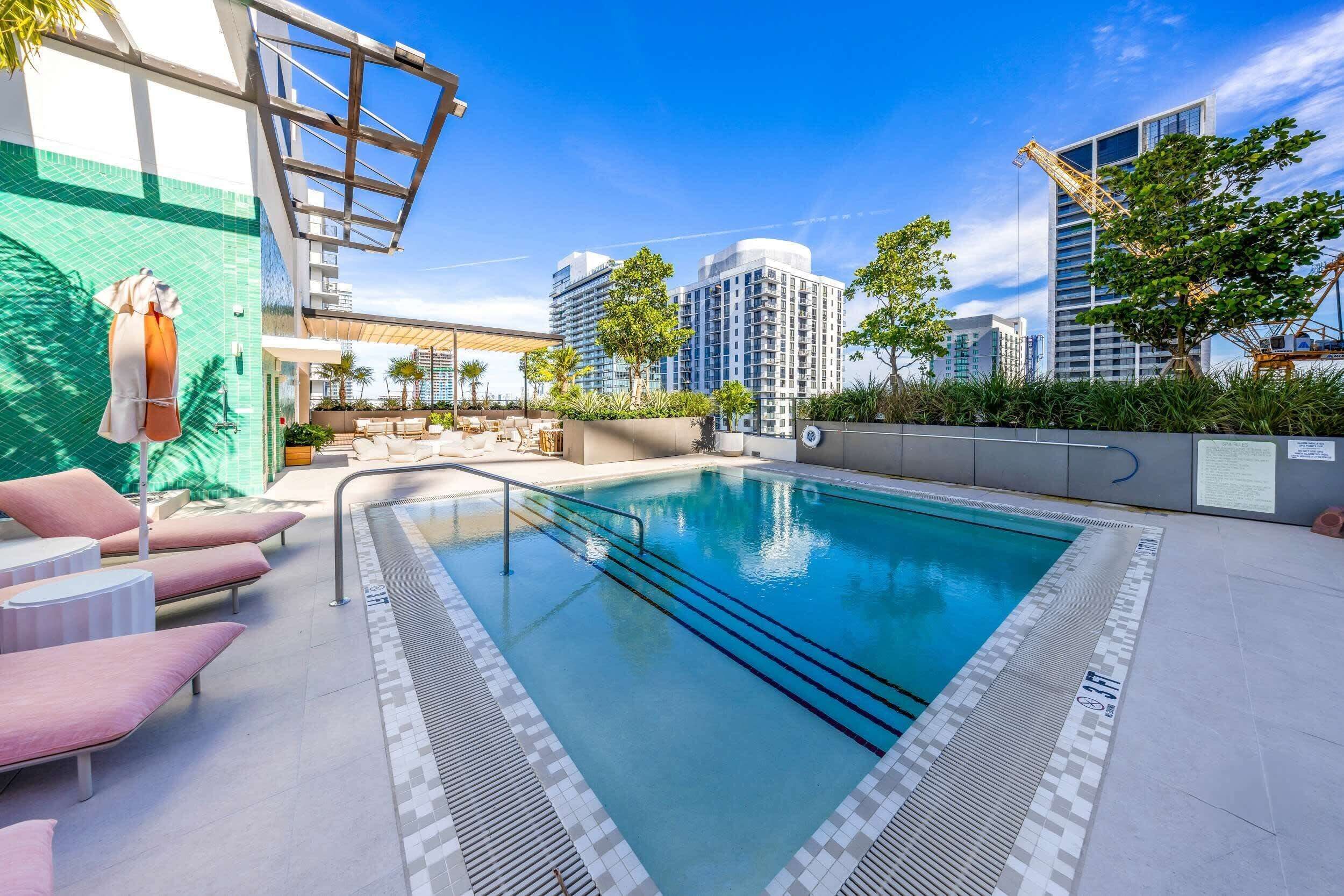 A rooftop pool with lounge chairs and a covered seating area, surrounded by high-rise buildings and greenery.