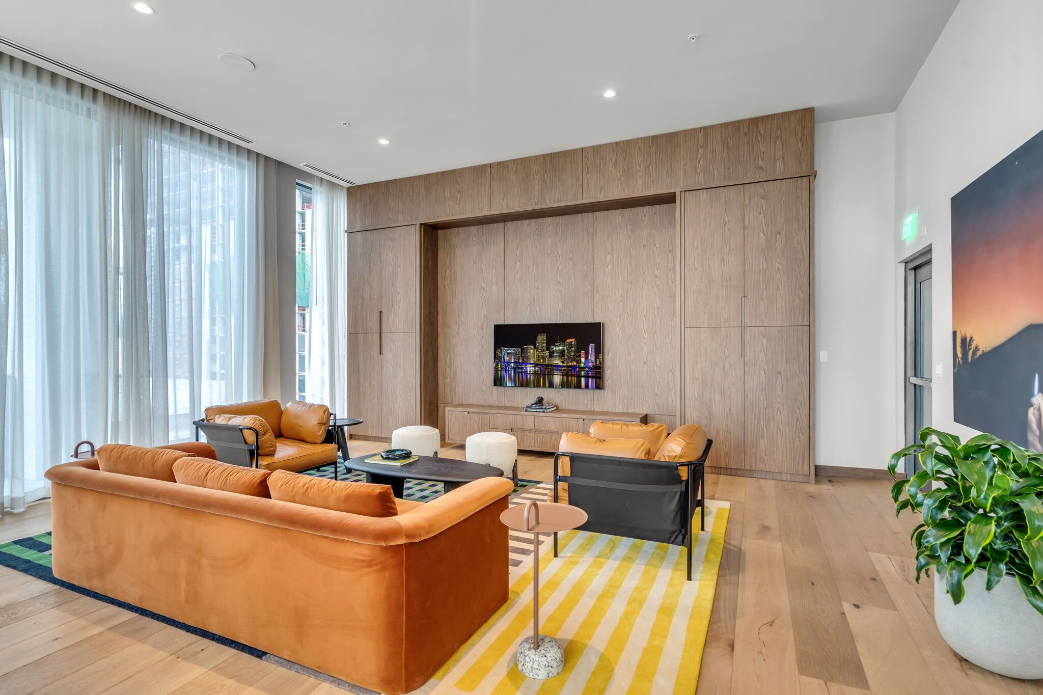 Uma sala de estar moderna com um sofá, poltronas e uma mesa de centro. Há uma televisão de tela plana na parede, um vaso de planta e uma janela grande com cortinas.