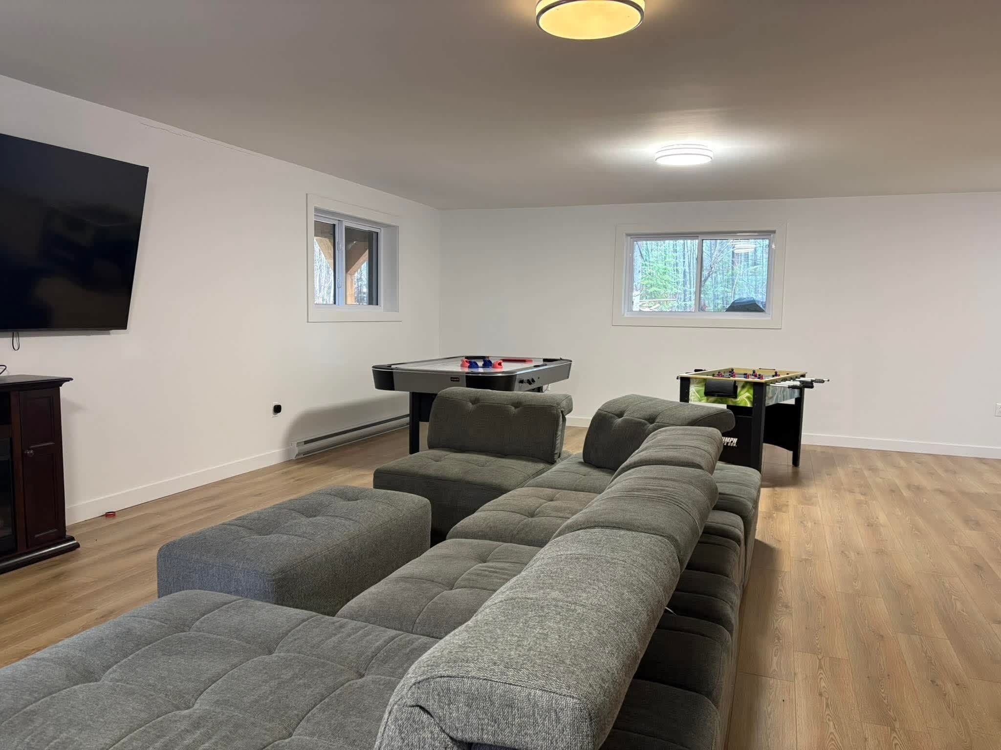 A living room with a sectional sofa, a flat-screen TV, a foosball table, and a window with a view of trees.