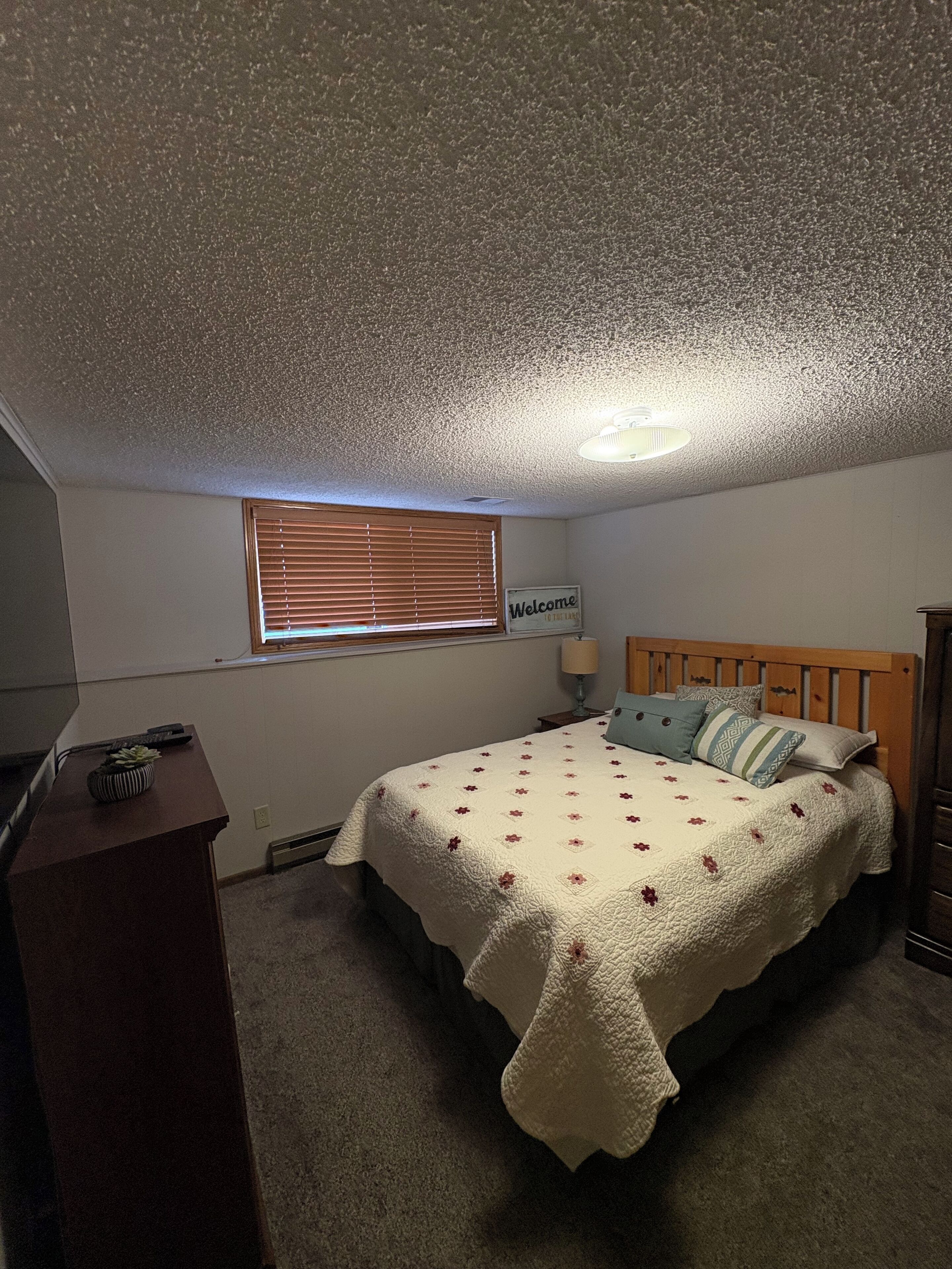 A bedroom with a bed, a desk, a window with blinds, a ceiling light, and a welcome sign.