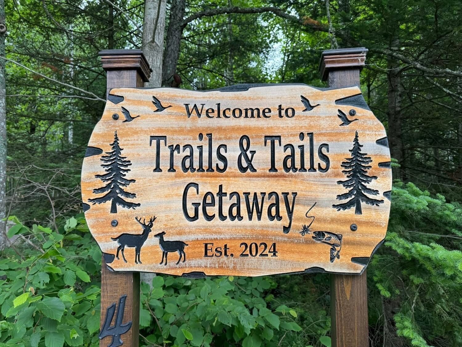 A wooden sign with the text "Welcome to Trails & Tails Getaway Est. 2024" surrounded by nature.
