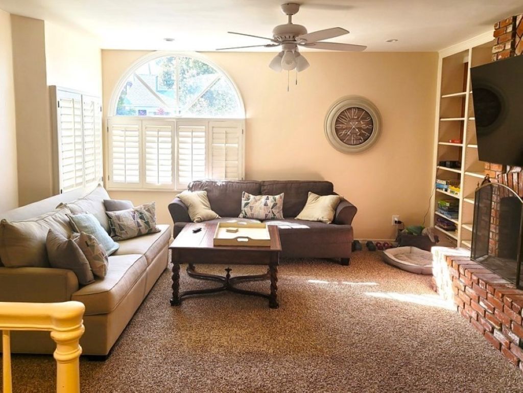 A living room with a brown sofa, a coffee table, a ceiling fan, a fireplace, and a large window with shutters.