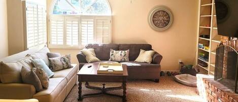A living room with a brown sofa, a coffee table, a ceiling fan, a fireplace, and a large window with shutters.