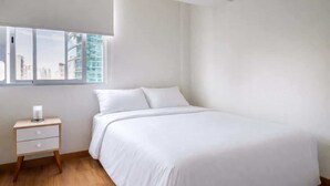 Ein schlicht eingerichtetes Schlafzimmer mit einem großen Bett, einem Nachttisch und einem Fenster mit Blick auf die Stadt.