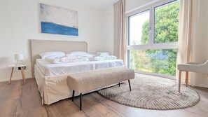 Ein Schlafzimmer mit Bett, einer Bank, einem Nachttisch, einem Sessel, einem Fenster und einem Bild an der Wand.
