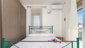A single bed with a metal frame, a bedside table, a window with curtains, and an air conditioning unit on the wall. - Apartment 'I Ciottoli Dello Ionio - Apt 18' with Sea View, Shared Terrace and Balcony (Mascali)