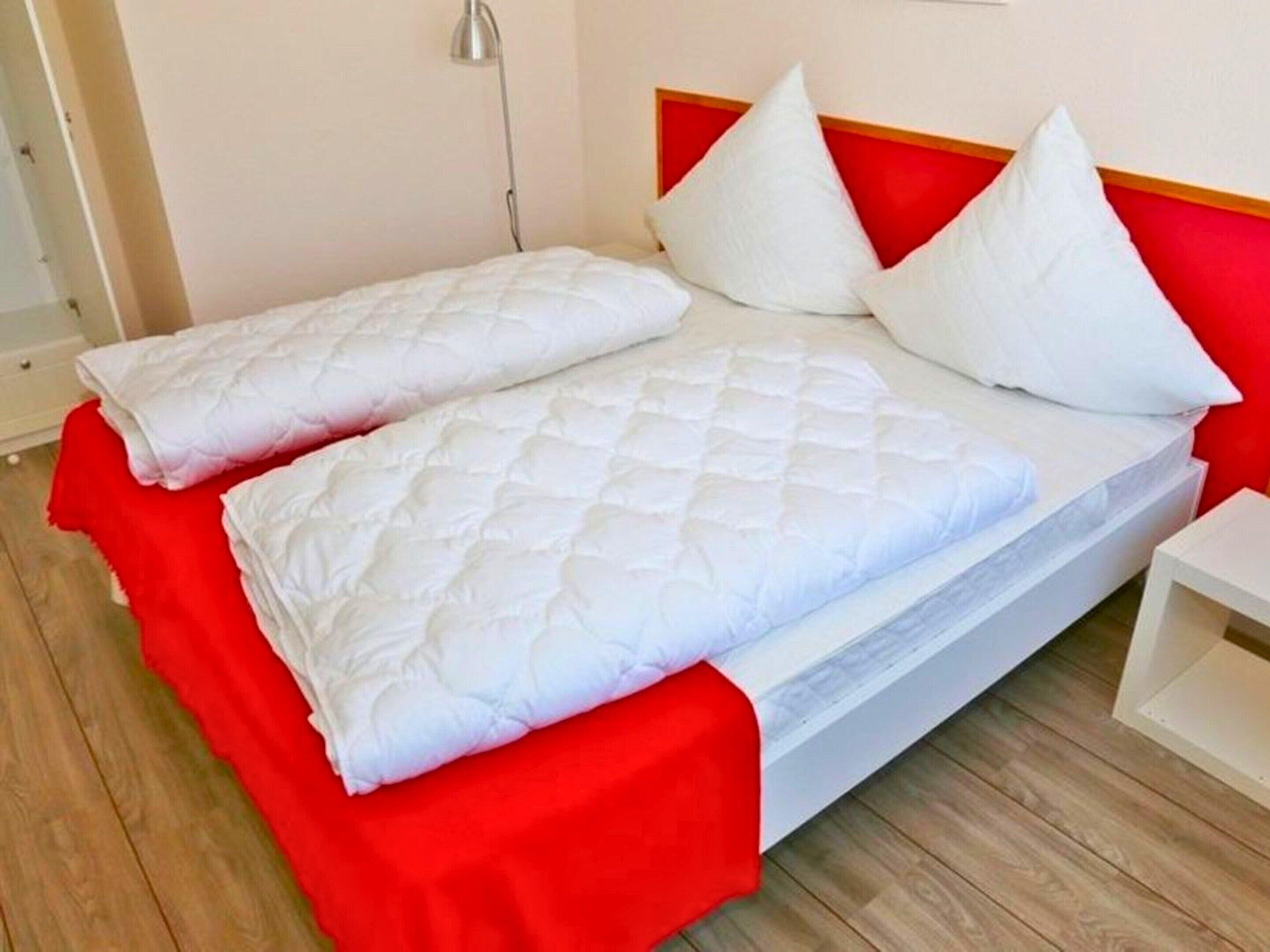 Una cama con ropa de cama blanca y detalles rojos, una mesita de noche y una lámpara.