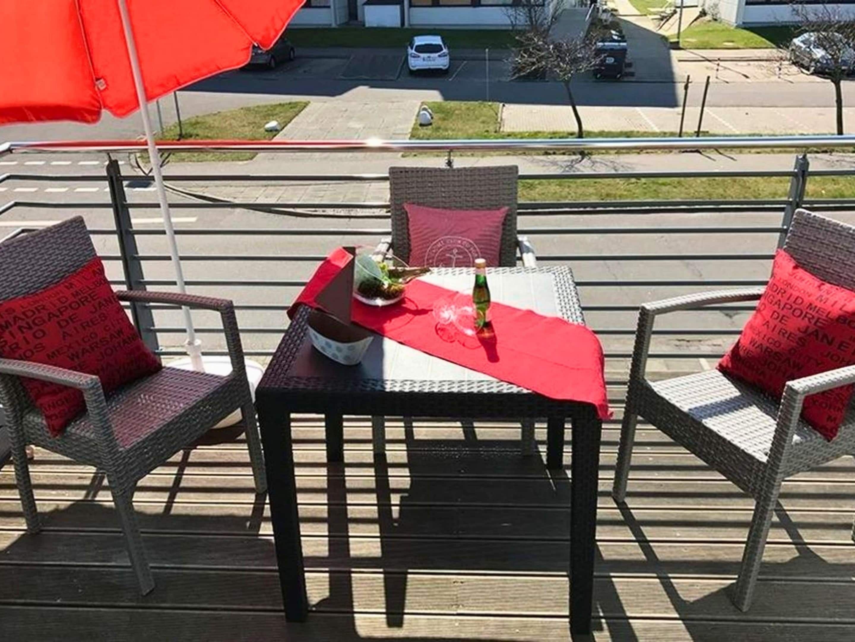 Une terrasse avec des chaises et une table en osier, recouverte d’une nappe rouge, sur laquelle se trouvent une bouteille et un bol.