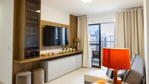 A modern living room with a flat-screen TV, a glass cabinet with decorative items, a sofa, a coffee table, and a view of the city through large windows. - Buenos Ayres #509 - Apt With Balcony by Carpediem (João Pessoa)