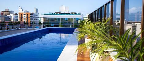 A rooftop pool with a wooden deck and potted plants, overlooking a cityscape with modern buildings.