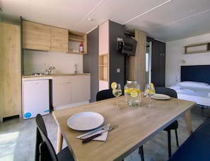 A compact kitchen with a refrigerator, sink, and wooden cabinets. A dining table with a plate, fork, and knife is set. A TV is mounted on the wall, and a bed is visible in the background. - Cozy Mobile Home in Cesenatico (Cesenatico)