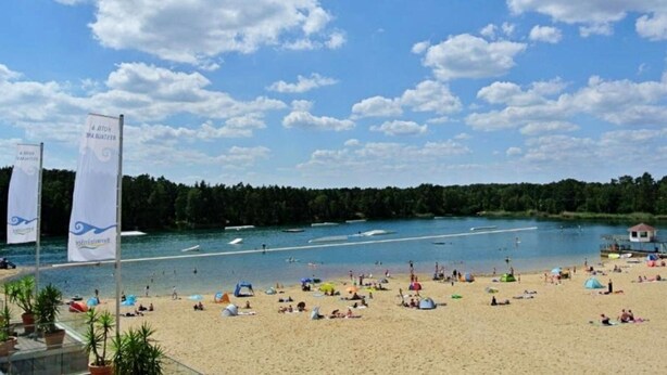A beach with a clear sky, people sunbathing, and a flag with text. - Bernsteinsee Homes and Ferien Beach House (Sassenburg)