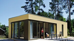 A modern, single-story building with large glass windows, a flat roof, and a patio area with outdoor furniture. - Bernsteinsee Homes and Ferien Beach House (Sassenburg)