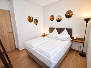 Een net opgemaakt bed met witte lakens, een houten hoofdbord en een nachtkastje met een lamp. Daarboven hangen drie decoratieve wandplaten.