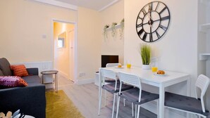 A modern living room with a sofa, a dining table with chairs, a large wall clock, and a small plant on the table. - 2-bedroom Bright London Home (London)