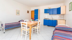 Une chambre avec deux lits simples, une table à manger en bois avec des chaises, un coin cuisine avec des placards bleus et un sol carrelé de couleur blanche.