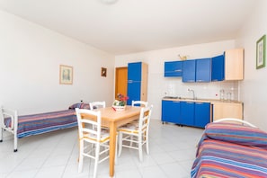 Una habitación con dos camas individuales, una mesa de comedor de madera con sillas, una zona de cocina con armarios azules y suelo de baldosas blancas.