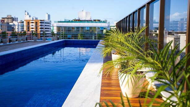 Una piscina sul tetto con un pontile in legno e piante in vaso, con vista su un paesaggio urbano di edifici moderni.