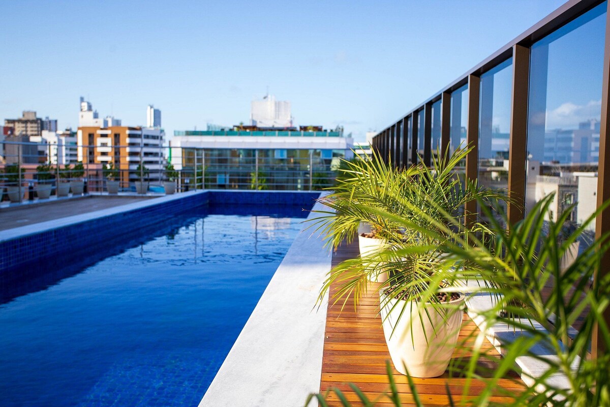 A rooftop pool with a wooden deck and potted plants, overlooking a cityscape with modern buildings.