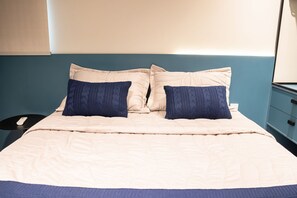 Un lit avec une couette beige et deux oreillers bleu foncé, une table de chevet avec un téléphone, et un mur bleu.