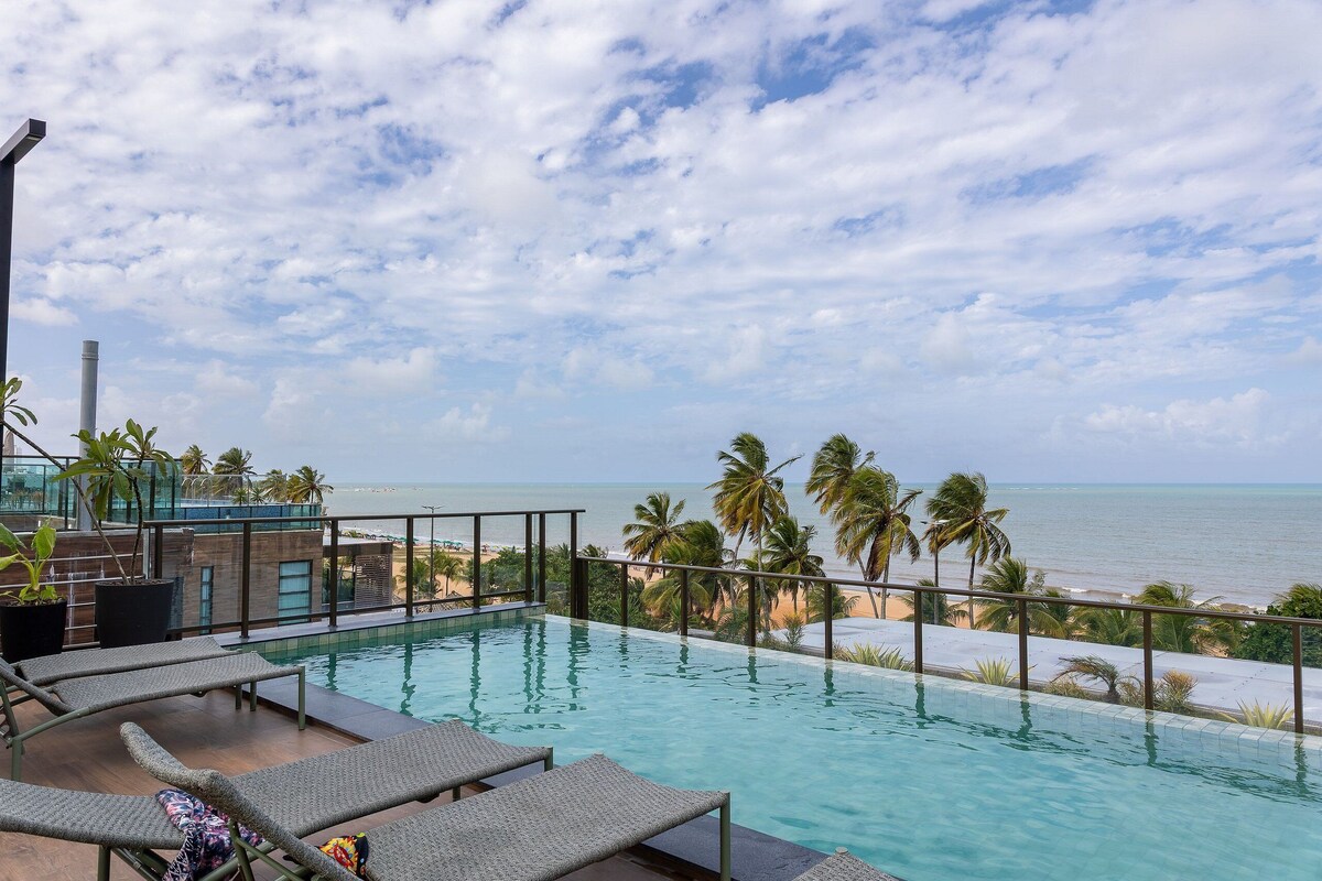 A rooftop infinity pool with lounge chairs, palm trees, and a view of the ocean.