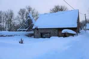 Une cabane en pierre, dotée d’un toit abrupt recouvert de neige, entourée d’un paysage enneigé et d’arbres sans feuilles.