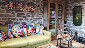 A cozy living room with a stone wall, a green sofa with colorful cow-themed cushions, a wooden cabinet with glass doors, and a framed golf poster. - Rustic Mont-dore Family Cottage (Mont-Dore)