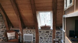 A cozy attic bedroom with a sloped wooden ceiling, a stone wall, a bed with a plaid blanket, a wicker basket on the window sill, and a television on a wooden stand. - Rustic Mont-dore Family Cottage (Mont-Dore)