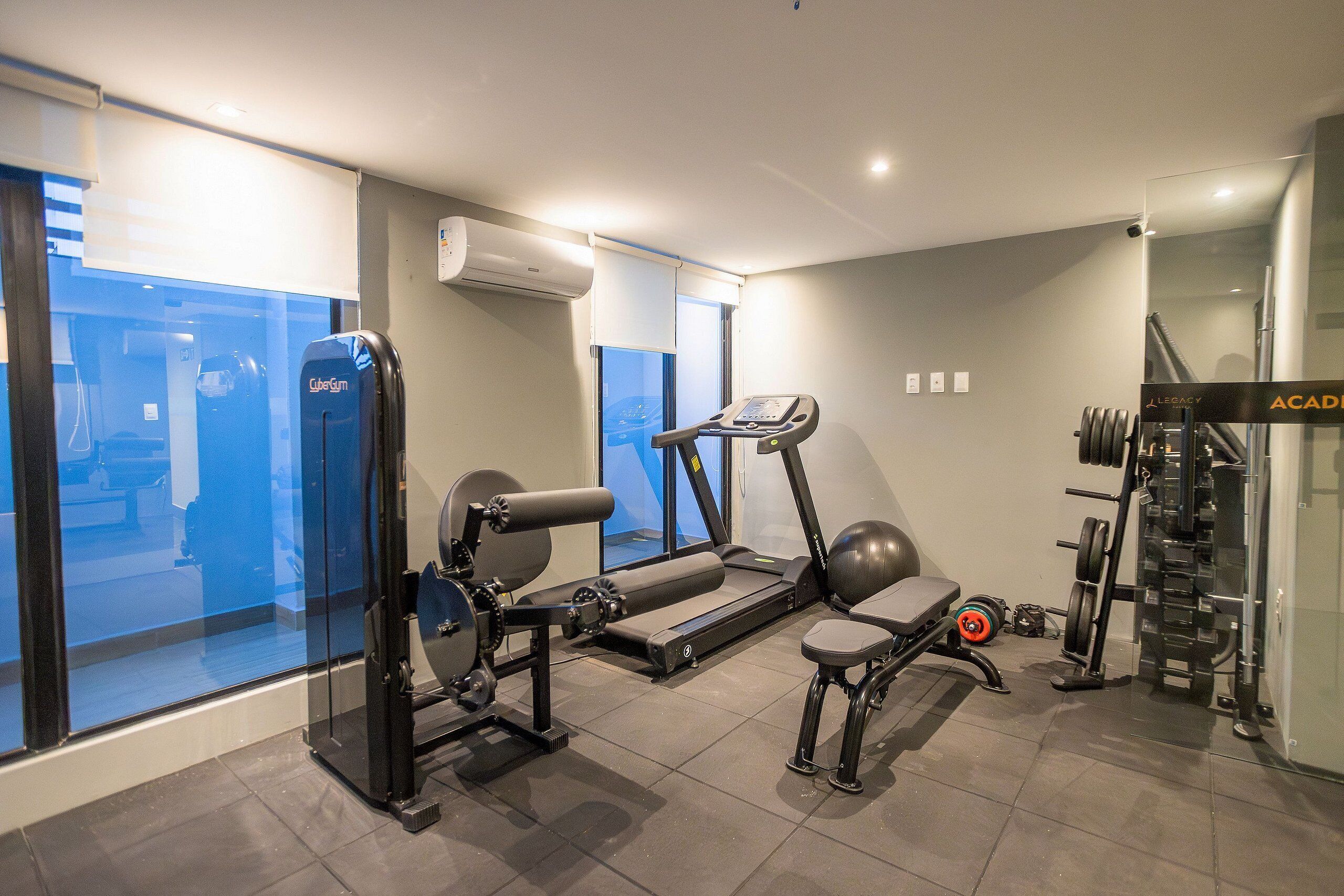 A modern gym with cardio equipment, weightlifting stations, and a large window offering a view of the city.