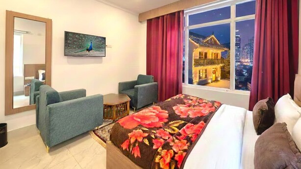 A hotel room with a large bed, two armchairs, a small table, a painting of a peacock, and a window with a city view at night. - Everglow Retreat White Tulip Guest House (Srinagar)